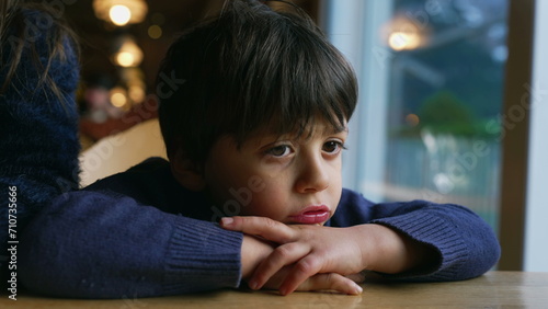 Pensive child feeling boredom at restaurant, leaning on table. Thoughtful little boy daydreaming, kid lost in thought