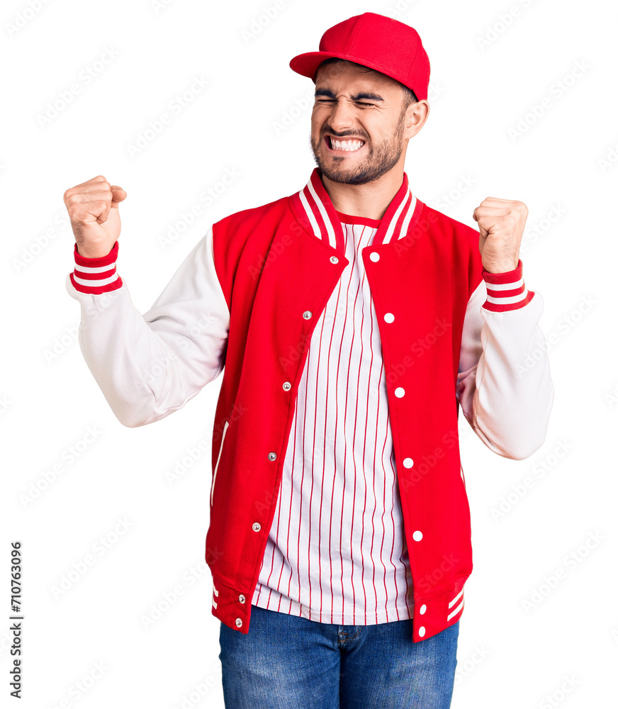 Young handsome man wearing baseball jacket and cap very happy and excited doing winner gesture with arms raised, smiling and screaming for success. celebration concept. Young handsome man wearing baseball jacket and cap very happy and excited doing winner gesture with arms raised, smiling and screaming for success. celebration concept.