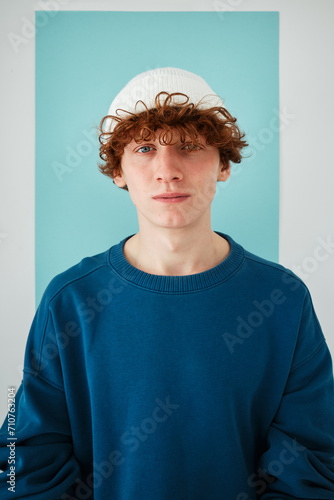 studio portrait on cyclorama of a young man with red curly hair in blue sweatshirt white beanie hat on blue paper background