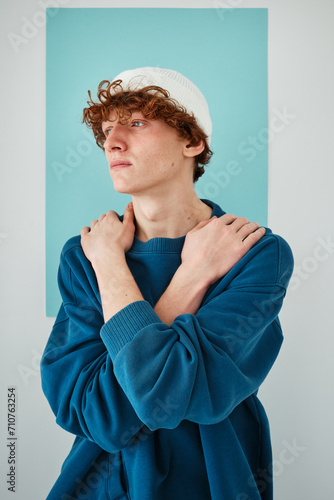 studio portrait on cyclorama of a young man with red curly hair in blue sweatshirt white beanie hat blue pants on blue paper background