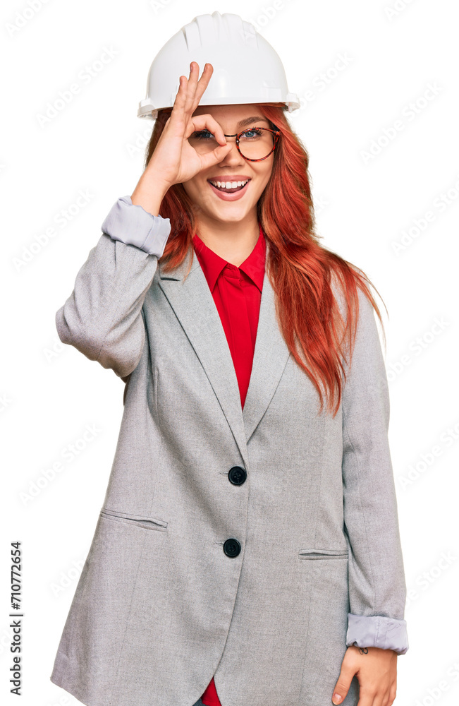Young redhead woman wearing architect hardhat doing ok gesture with hand smiling, eye looking through fingers with happy face. Young redhead woman wearing architect hardhat doing ok gesture with hand smiling, eye looking through fingers with happy face.