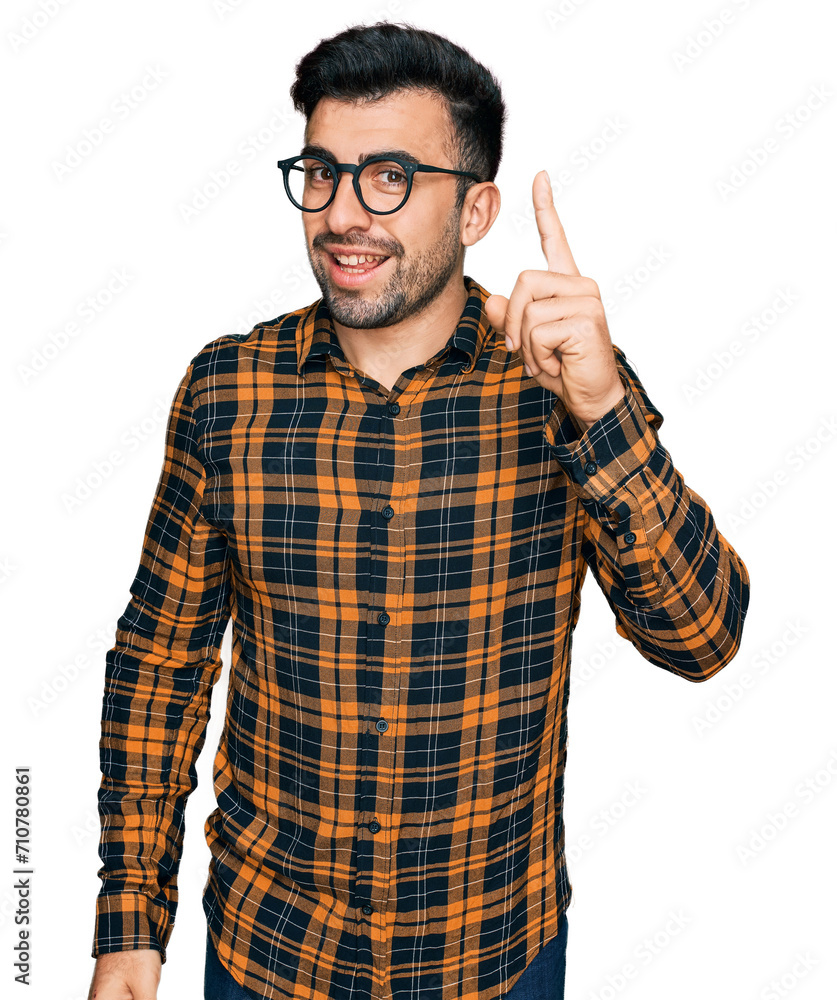 Hispanic man with beard wearing casual clothes and glasses pointing finger up with successful idea. exited and happy. number one. Hispanic man with beard wearing casual clothes and glasses pointing finger up with successful idea. exited and happy. number one.