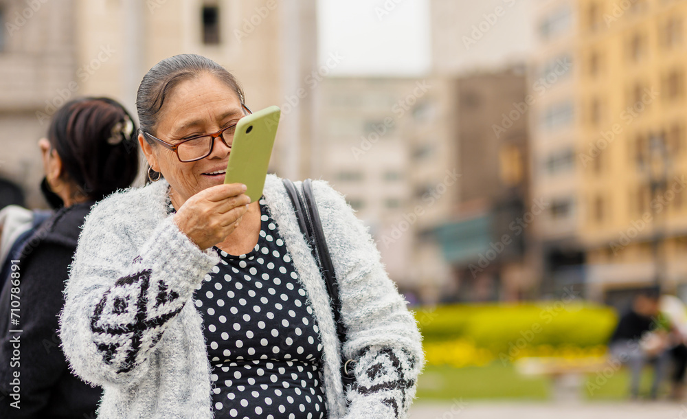 Fototapeta premium Latina adult woman, with a happy expression, in the city using her smartphone, sending audio messages and typing. Copyspace......