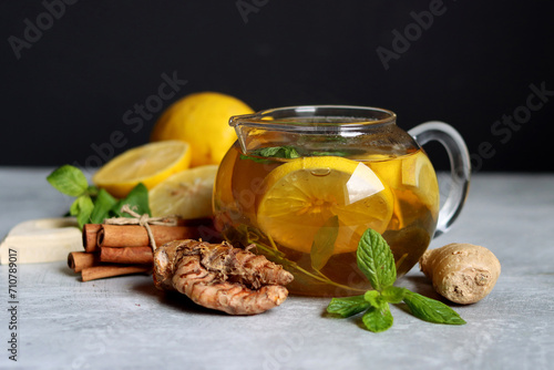 Teapot with hot tea, ginger, turmeric root, cinnamon sticks, lemon and mint leaves on grey background with copy space. Anti inflammatory winter drink recipe. 