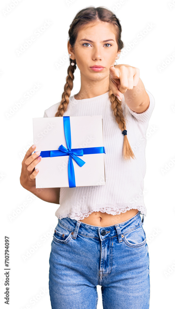 Beautiful caucasian woman with blonde hair holding gift pointing with finger to the camera and to you, confident gesture looking serious