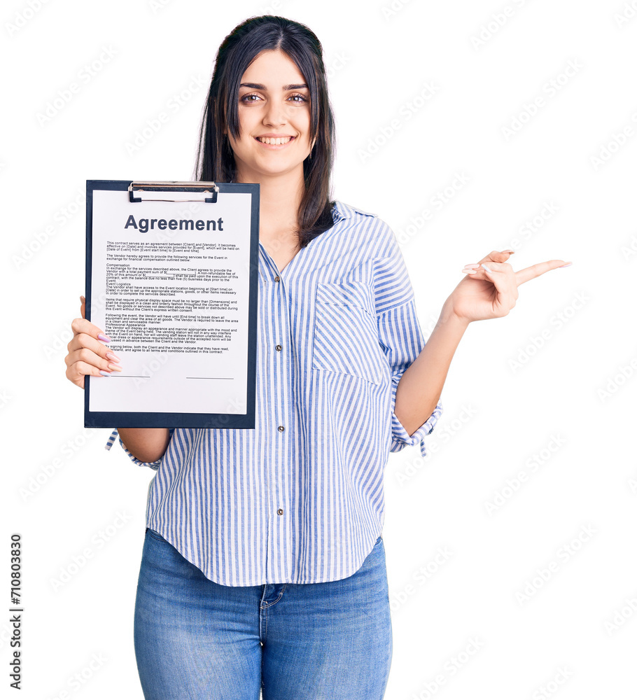 Young beautiful girl holding clipboard with agreement document smiling happy pointing with hand and finger to the side Young beautiful girl holding clipboard with agreement document smiling happy pointing with hand and finger to the side