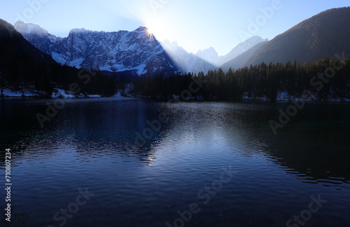 sun peeking behind the mountains and the reflection of the mountains on the beautiful alpine lake in winter without people