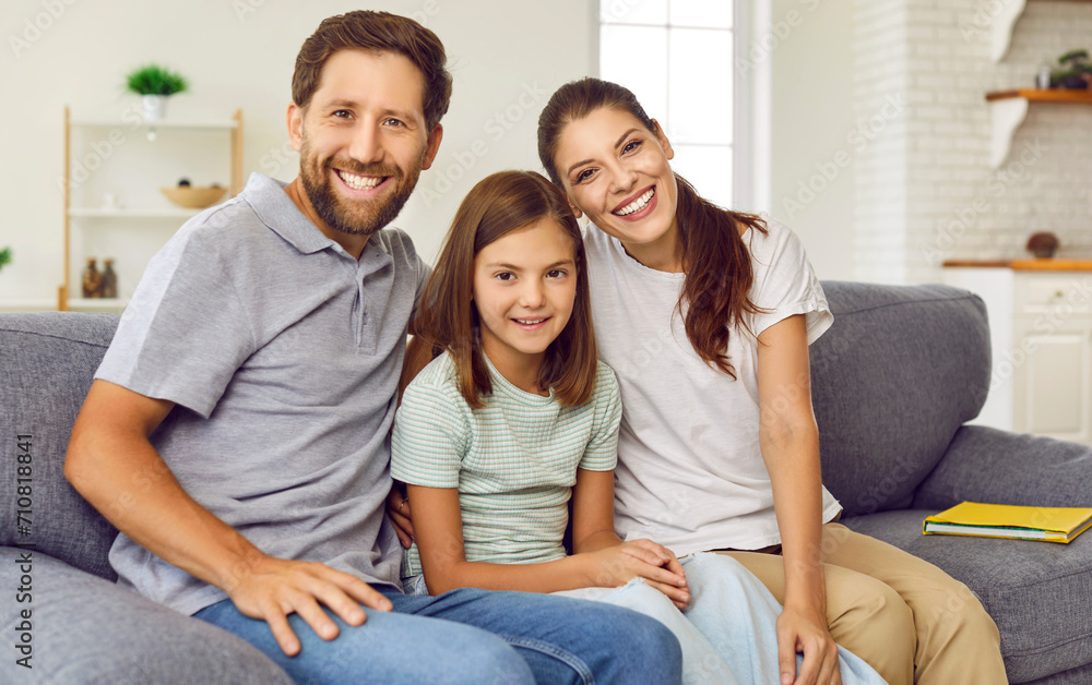 Portrait of smiling young happy family of three with daughter sitting on the sofa at home and looking cheerful at camera. Child girl sitting with parents. Love, care, family time and leisure concept. Portrait of smiling young happy family of three with daughter sitting on the sofa at home and looking cheerful at camera. Child girl sitting with parents. Love, care, family time and leisure concept.