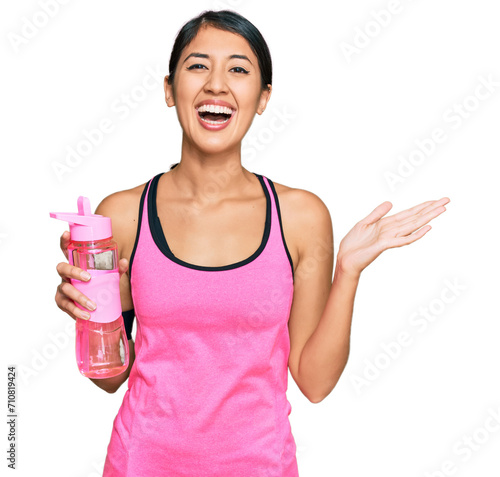 Beautiful asian young sport woman wearing sportswear drinking bottle of water celebrating victory with happy smile and winner expression with raised hands