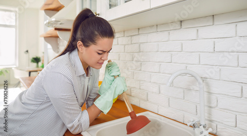 Woman having a problem with the sink. Young woman doing work about the house and trying to unblock the food remains clogged in the kitchen sink drain with a plunger. Housework concept photo