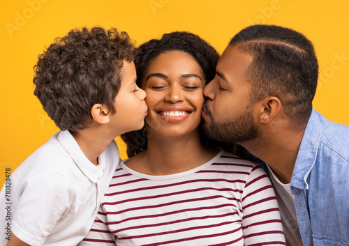 Adorable son and husband giving kisses on cheeks of smiling black mother