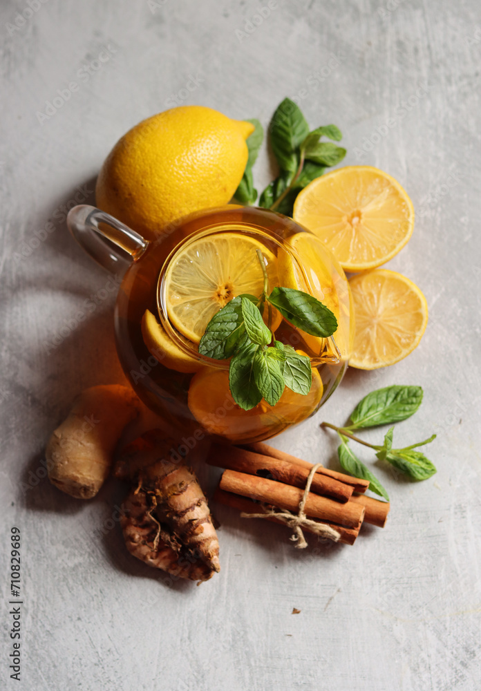 Teapot with hot tea, ginger, turmeric root, cinnamon sticks, lemon and mint leaves on grey background with copy space. Anti inflammatory winter drink recipe. Teapot with hot tea, ginger, turmeric root, cinnamon sticks, lemon and mint leaves on grey background with copy space. Anti inflammatory winter drink recipe.