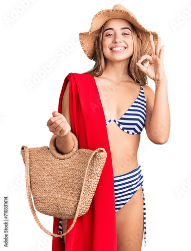 Young beautiful blonde woman wearing bikini and hat holding summer wicker handbag doing ok sign with fingers  smiling friendly gesturing excellent symbol