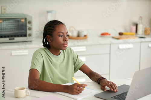 Black teenage girl studying at home, reading article on laptop and writing in copybook