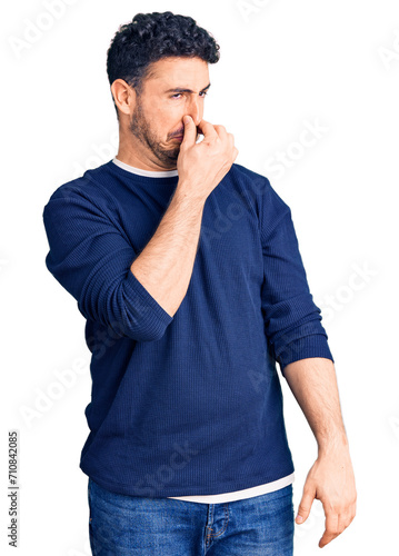 Young hispanic man wearing casual clothes smelling something stinky and disgusting, intolerable smell, holding breath with fingers on nose. bad smell