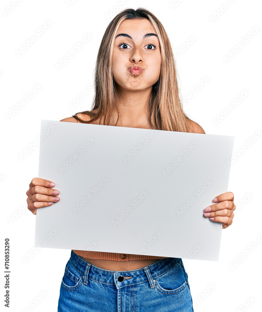 Hispanic young woman holding blank empty banner puffing cheeks with funny face. mouth inflated with air, catching air. Hispanic young woman holding blank empty banner puffing cheeks with funny face. mouth inflated with air, catching air.
