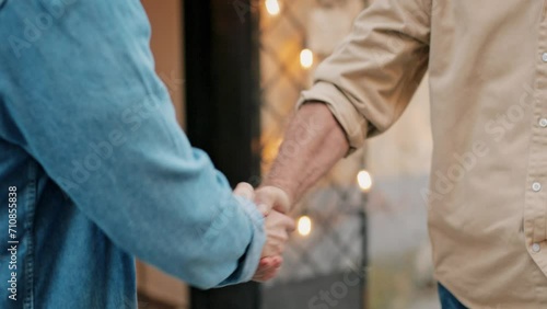 Camera filming firm handshake of two men. People greeting each other in day light in middle of street. Shaking hands in front of restaurant or cafe. Blurred building in background.