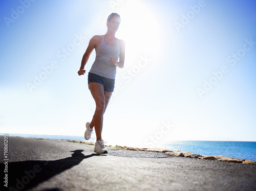 Woman, fitness and running on beach for workout, exercise or outdoor training on a sunny day. Female person, athlete or runner in race, sprint or sports marathon on asphalt, road by the ocean coast © Miko/peopleimages.com