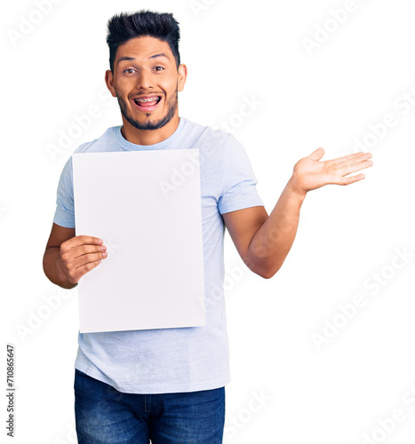 Handsome latin american young man holding cardboard banner with blank space celebrating victory with happy smile and winner expression with raised hands