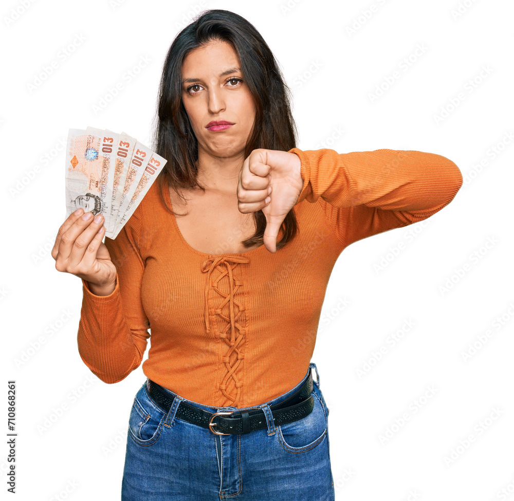 Beautiful hispanic woman holding 10 united kingdom pounds banknotes with angry face, negative sign showing dislike with thumbs down, rejection concept Beautiful hispanic woman holding 10 united kingdom pounds banknotes with angry face, negative sign showing dislike with thumbs down, rejection concept
