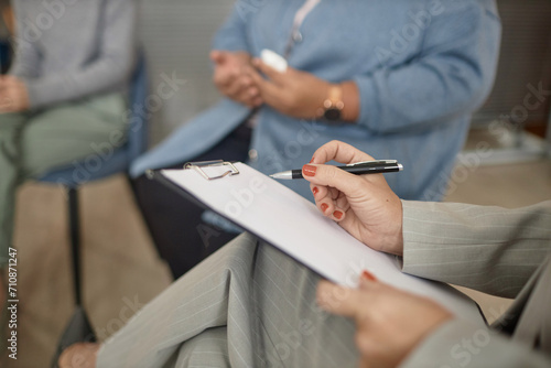 Closeup of unrecognizable female therapist holding clipboard and pen taking notes in support group session, copy space
