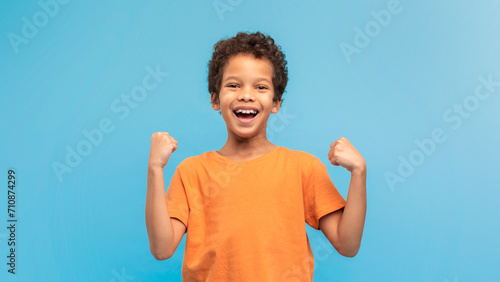 Foto Exuberant boy in orange shirt, fists raised, blue background