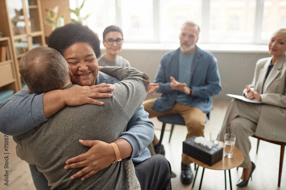 © Seventyfour - Portrait of Black senior woman embracing friend in group therapy session and smiling happily celebrating recovery, copy space