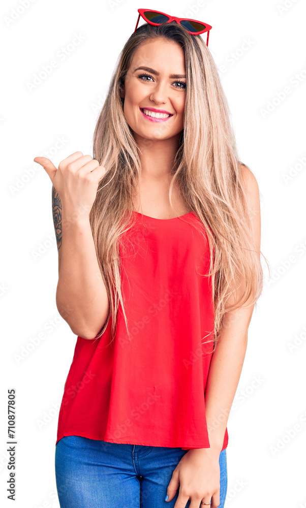 Young beautiful blonde woman wearing sleeveless t-shirt and sunglasses smiling with happy face looking and pointing to the side with thumb up. Young beautiful blonde woman wearing sleeveless t-shirt and sunglasses smiling with happy face looking and pointing to the side with thumb up.