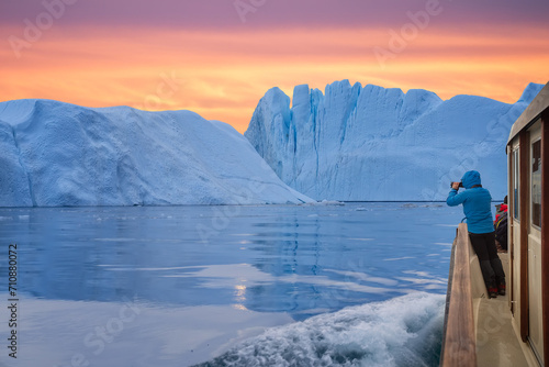 Tourist in a boat in front of an iceberg taking photos at midnight sun sunset. Tourist man explorer watching a large iceberg melting in the arctic sea from the coast of Ilulissat. Greenland