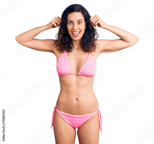 Young beautiful hispanic woman wearing bikini smiling pulling ears with fingers, funny gesture. audition problem
