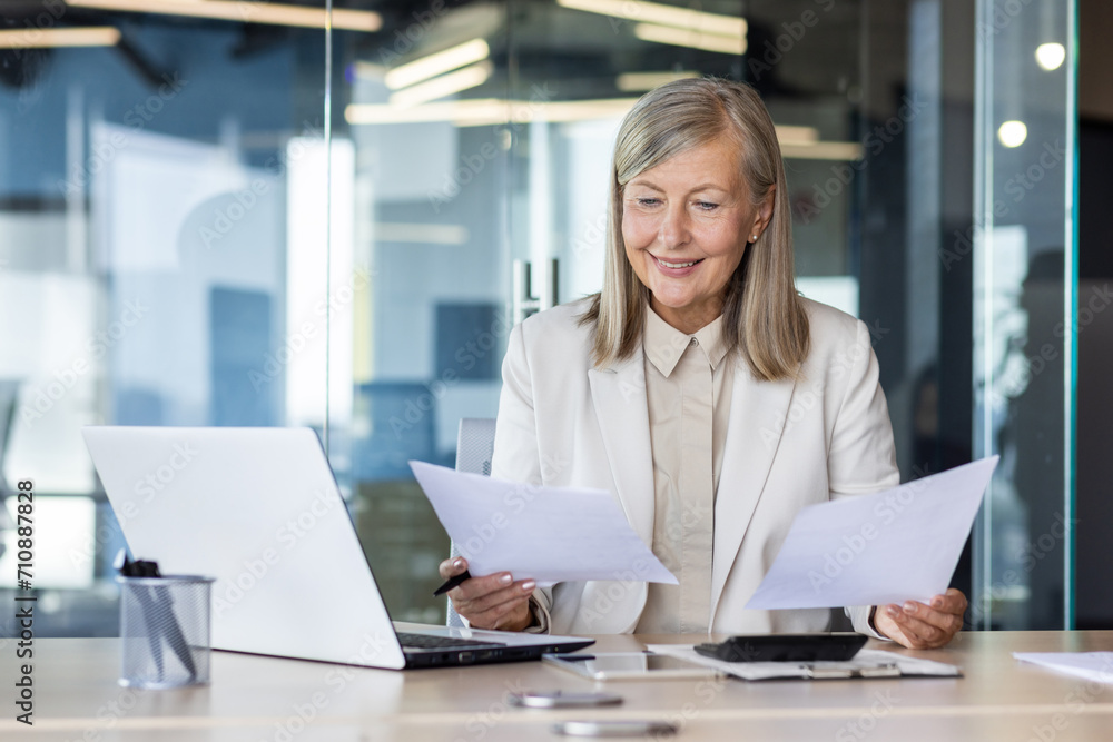 Smiling senior business woman working in office at desk, holding documents in hands, reviewing deal and invoices. Smiling senior business woman working in office at desk, holding documents in hands, reviewing deal and invoices.