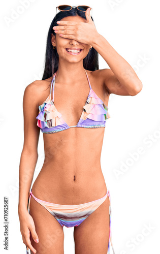 Young beautiful latin girl wearing bikini and sunglasses smiling and laughing with hand on face covering eyes for surprise. blind concept.