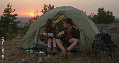 A young man and his girlfriend have dinner and make delicious coffee near a tent in the mountains after climbing. A couple in love rests near a tent at sunset.