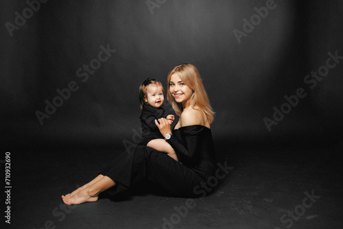 Portrait of a young mother holding her baby daughter in her arms, hugging and smiling while sitting in a studio on a black background