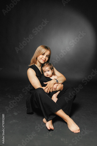 Happy family, motherhood. Beautiful happy mother hugs her little newborn daughter and looks at the camera. Studio portrait on black background