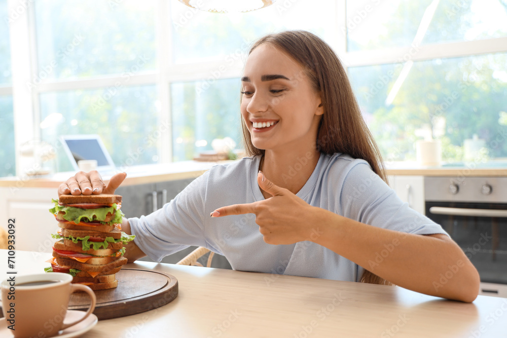 Beautiful young woman pointing at big sandwich on table in kitchen