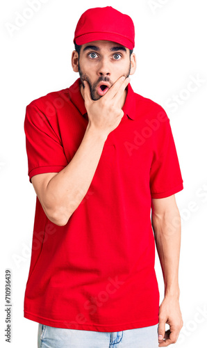 Young handsome man with beard wearing delivery uniform looking fascinated with disbelief, surprise and amazed expression with hands on chin