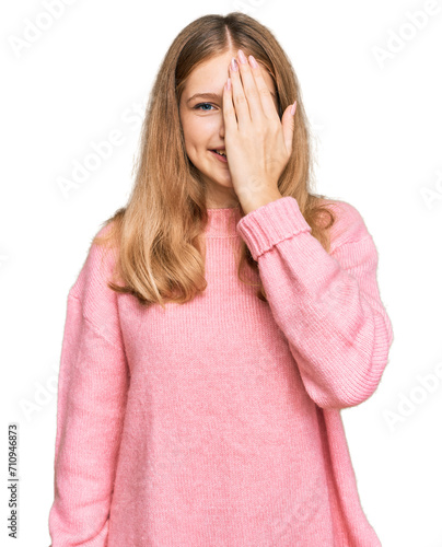 Beautiful young caucasian girl wearing casual winter sweater covering one eye with hand, confident smile on face and surprise emotion.