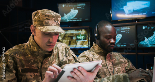 African American young man in headset typing on laptop computer and Caucasian soldier tapping on tablet device in control room. Multiethnic army officers in uniforms working together in office. photo