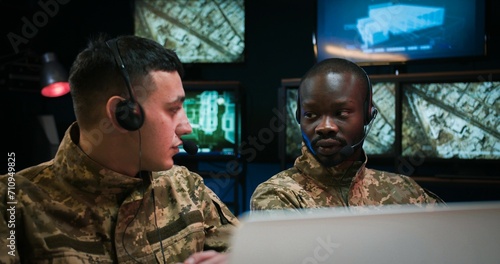 Multiethnic male military dispatchers colleagues in headsets sitting in monitoring room with screens and controlling war operation in conflict zone. Two mixed-races men cooperating in control room.