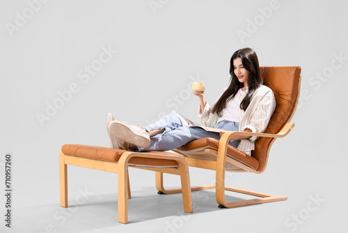 Beautiful young woman with cup of tea reading magazine in armchair on grey background