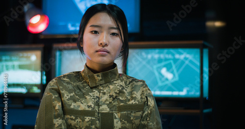 Portrait of pretty Asian female soldier standing in monitoring room and looking staright at camera. Attractive woman in militarian uniform working in troops. Girl in army concept. photo