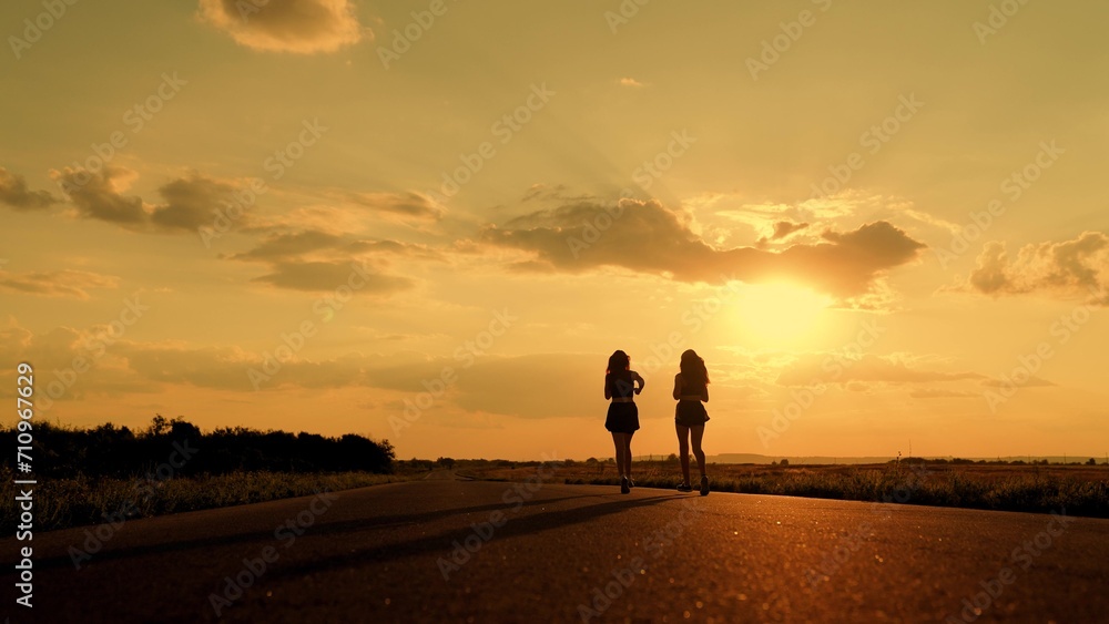 Athletic young women running along an asphalt road at sunset, teamwork, healthy fitness lifestyle. People run in sun together. Girls are training, running along road, teamwork of athletes. Slow motion
