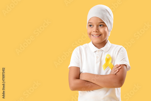 Cute little African-American boy after chemotherapy with yellow ribbon on color background. Childhood cancer awareness concept