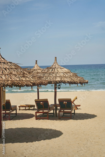 Fototapeta Naklejka Na Ścianę i Meble -  Straw sunshades and sunbeds on the empty pebble beach with sea in the background. Deserted beach with rattan sun loungers and umbrellas