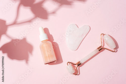 Cosmetic serum bottle, face roller and gua sha stone with orchid flower shadow on pink background. Sharp shadows. Home spa concept. Top view, flat lay