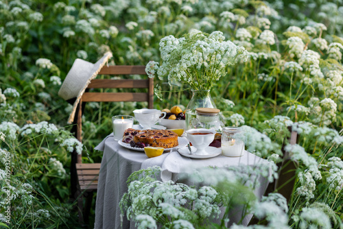 Tea time with sweet homemade cherry cake and herbal beverage in summer garden outdoors. Table covered with linen tablecloth, white porcelain cups. English style