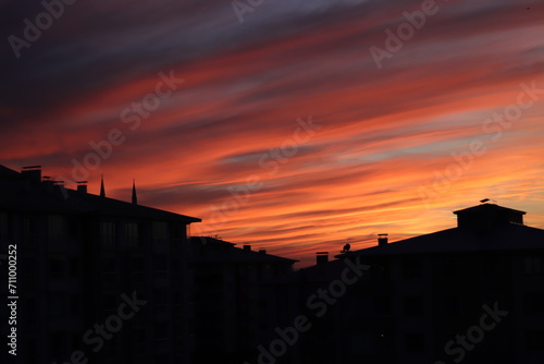 A dramatic sunset with radiant hues of orange and red streaking across the sky, silhouetting the rooftops of urban buildings. photo