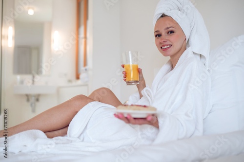 Cute young woman in bath robe having breakfast and feeling enjoyed