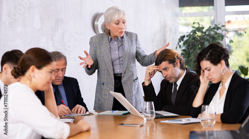 Irritated elderly female boss reprimanding upset subordinates sitting at table in office meeting room, expressing dissatisfaction with work photo